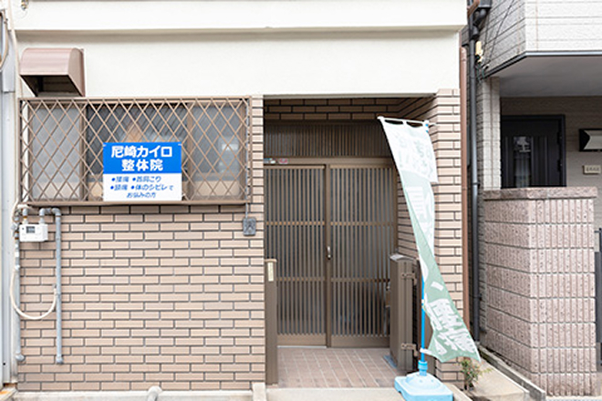 尼崎カイロ整体院の入り口外観。茶色のレンガ調の壁面に「尼崎カイロ整体院」と書かれた青い看板が掲げられており、格子状の引き戸の前に「腰痛」と書かれたのぼり旗が立っています