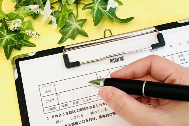 バインダーに挟まれた整体院の問診票に、ペンを持って記入している手元のアップ。背景の黄色いデスクの上には、緑色のアイビーのフェイクグリーンが添えられています。