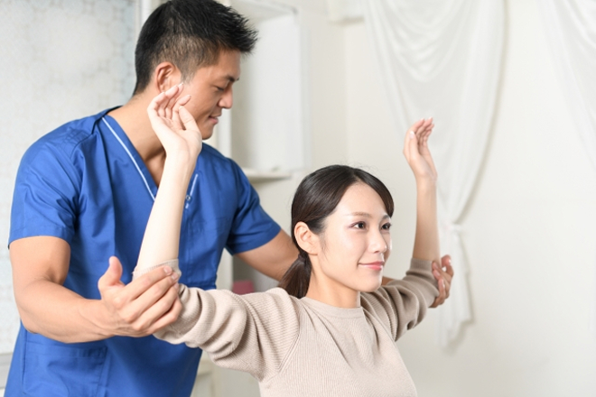 男性整体師が、両腕を上にあげた女性患者様の肘を後ろから支えながら、肩関節の可動域や筋肉の張りを優しく確認している施術・検査の様子。
