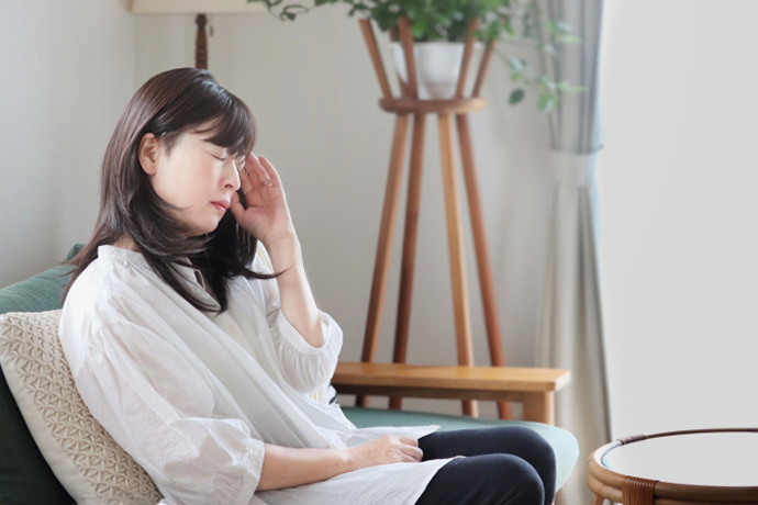 基本パターン（推奨）: リビングのソファにもたれかかり、目を閉じて辛そうな表情で左の側頭部を押さえる女性の画像。頭痛やめまいに悩む様子