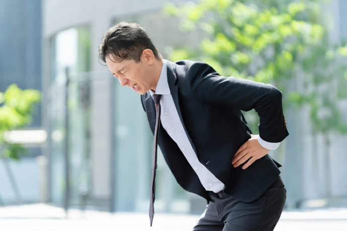 屋外でスーツを着たビジネスマンの男性が、顔をしかめて激しい腰の痛みに耐えながら、左腰の後ろに手を当てて前かがみになっている画像。