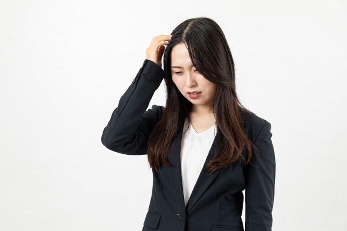 白背景の前で、黒いスーツを着たロングヘアの女性が、困ったような辛そうな表情で右手で頭をかきむしるように押さえている上半身の画像。