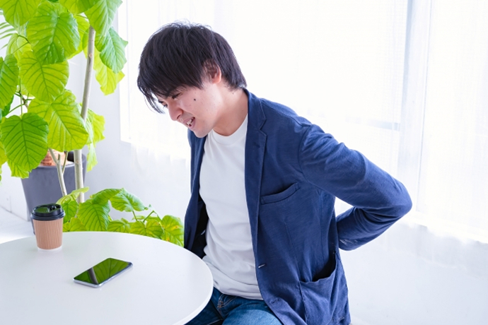 明るい室内で、ネイビーのジャケットを着た男性が、椅子に座りながら顔をしかめて両手で腰の後ろを押さえている、腰痛に悩む様子を捉えた画像。