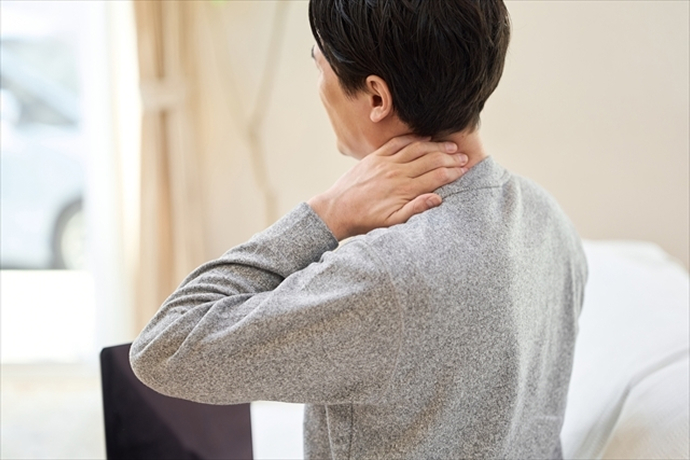 室内でグレーのトップスを着た男性が、左手で右の首筋から肩にかけてを押さえ、辛そうに少し頭を傾けている斜め後ろからの画像。