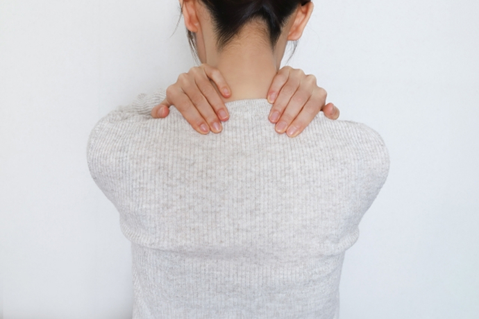 白い背景の前で、グレーのニットを着た女性が後ろを向き、両手で首の付け根から肩にかけてを揉んでいる様子。肩こりや首の痛みのイメージ。